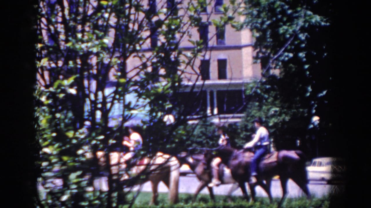 In 1958 French Lick, Indiana, horseback riders move past trees with a large building and mid-century cars in the background.
