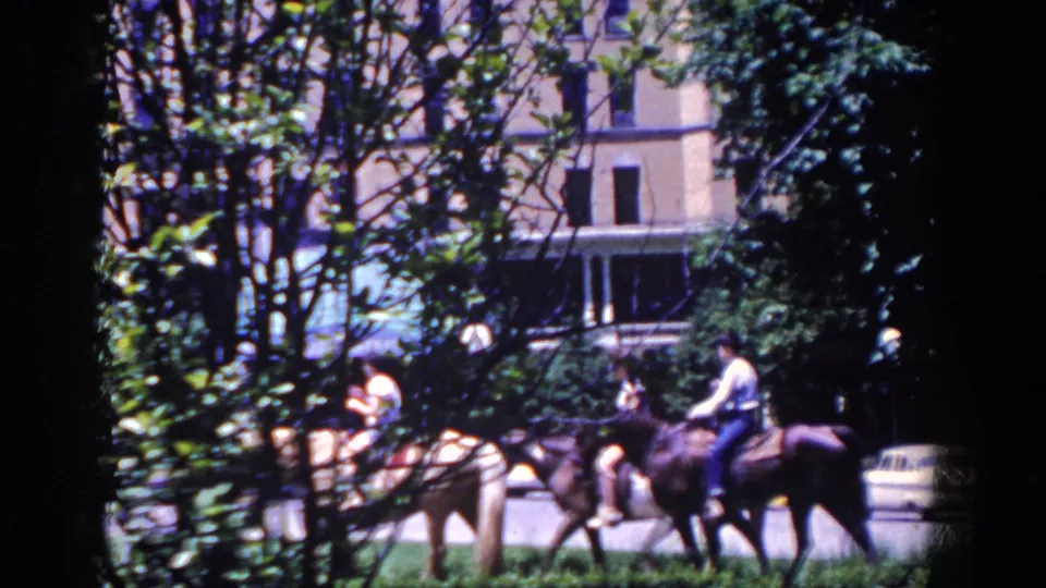 In 1958 French Lick, Indiana, horseback riders move past trees with a large building and mid-century cars in the background.