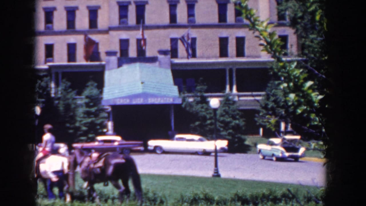 Riders on horses move across a lawn edge with parked cars and a large building behind them.