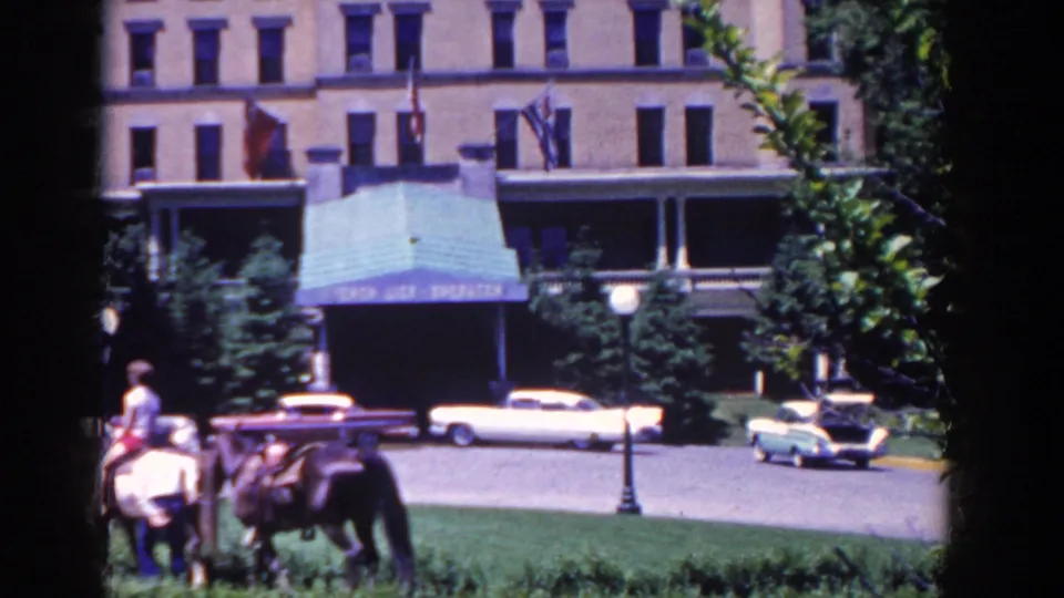 Horseback riders pass in front of a large French Lick building, with 1950s cars and shaded landscaping visible behind them.