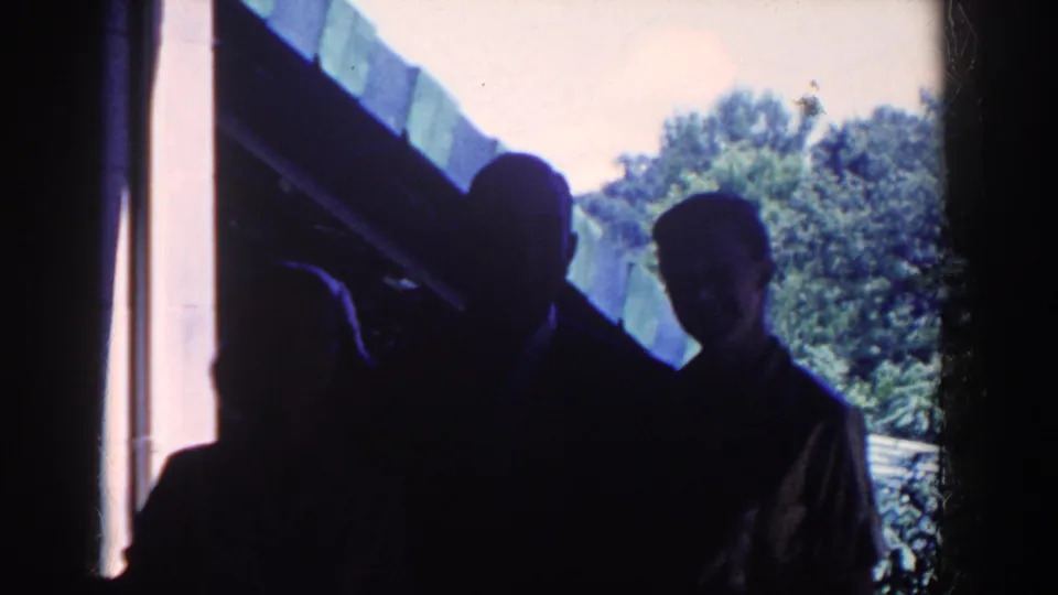 The silhouetted group remains clustered at the house entrance, with one adult leaning forward against the bright outdoor background.