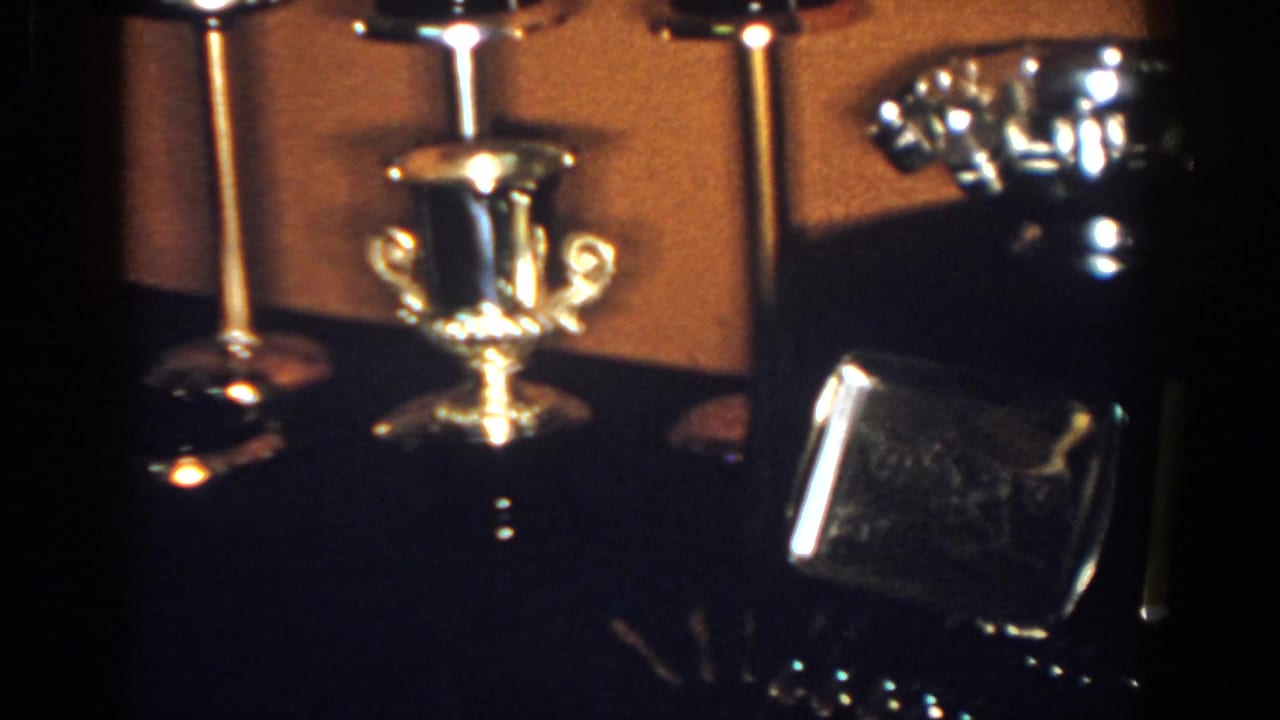 The camera holds on the metalware arrangement, with the handled cup centered between candlesticks and a polished rectangular case nearby.