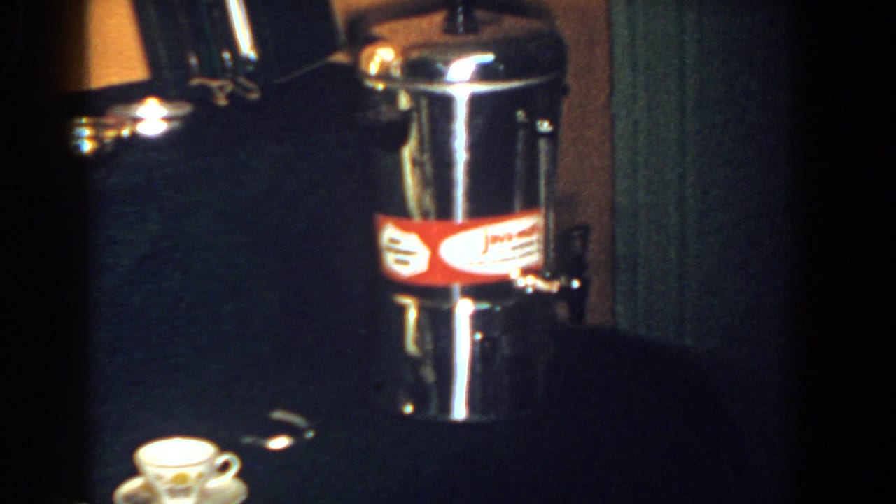 Metal coffee percolator with red label beside a small cup on a dark display surface.