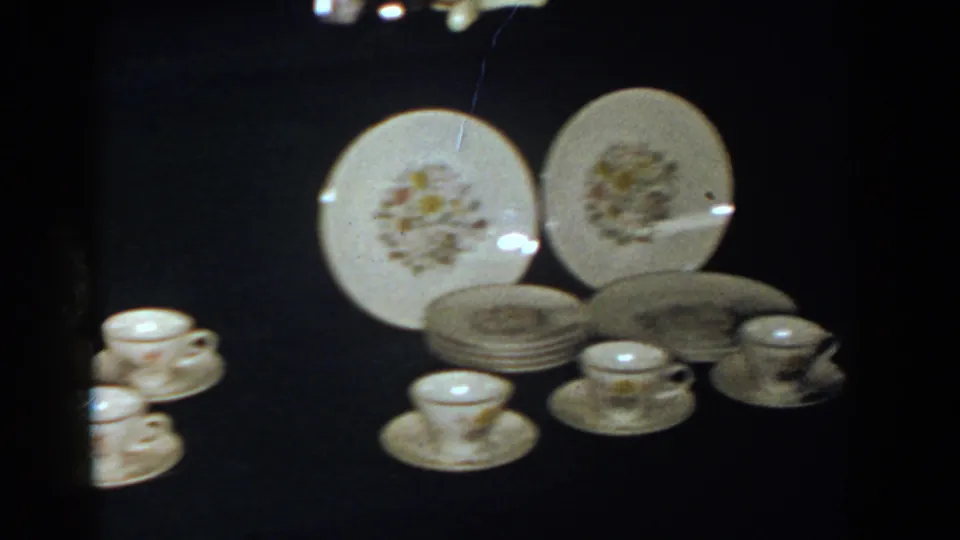 1958 French Lick frame showing a fuller view of floral plates, stacked dishes, cups, and saucers on a dark display surface.