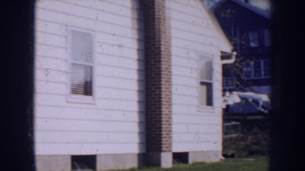 1956 Nebraska home-movie frame showing a white-sided house, brick chimney, small window, lawn, and neighboring building edge.