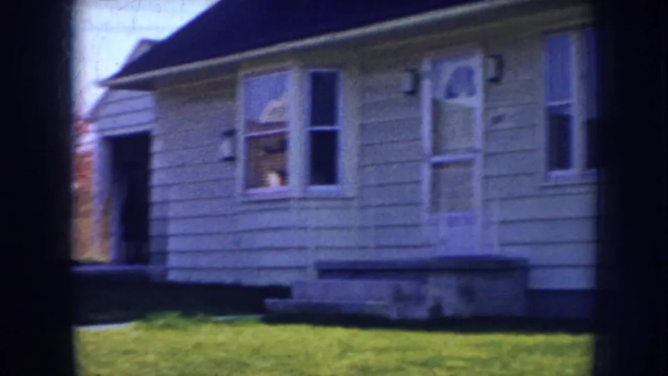 1950s home-movie view of a Nebraska house front with porch steps, lawn, window openings, siding, and a dark doorway at left.