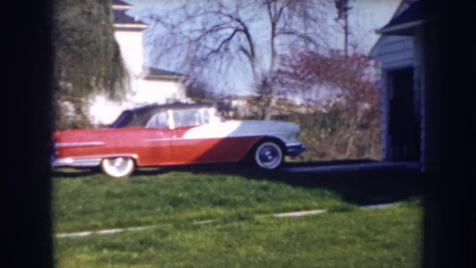 A red-and-white 1950s convertible is parked on a grassy driveway area between suburban houses in Nebraska.