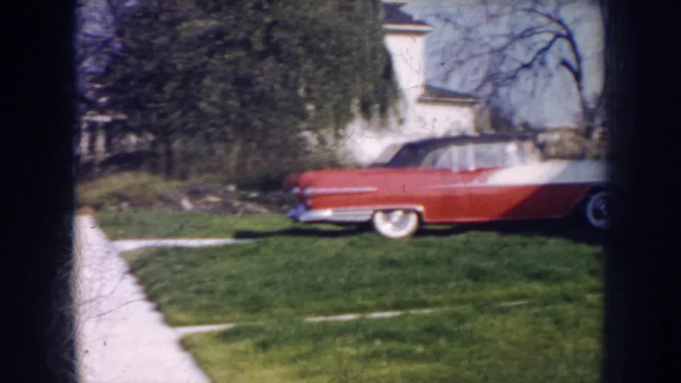 1956 Nebraska home-movie frame of a red-and-white convertible on a grassy front yard near a light-colored house.