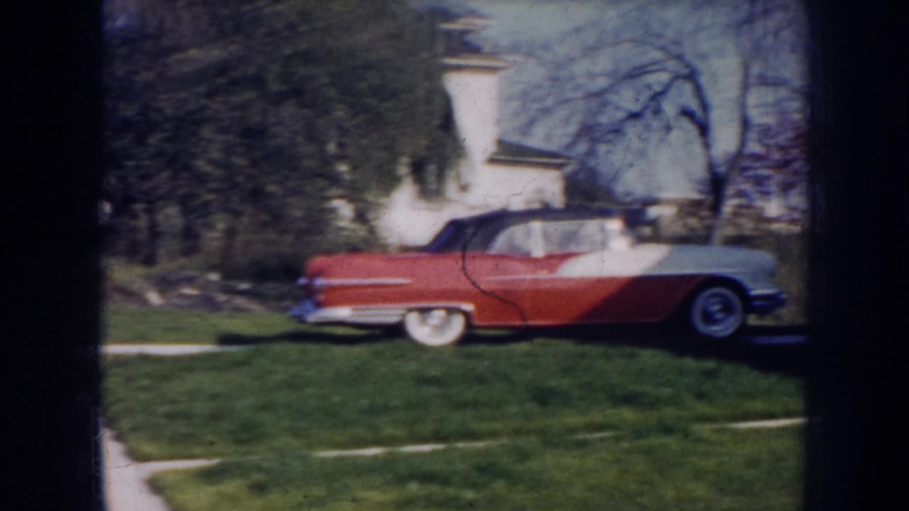 1956 Nebraska frame showing the side of a red-and-white convertible in a residential yard with bare trees behind it.