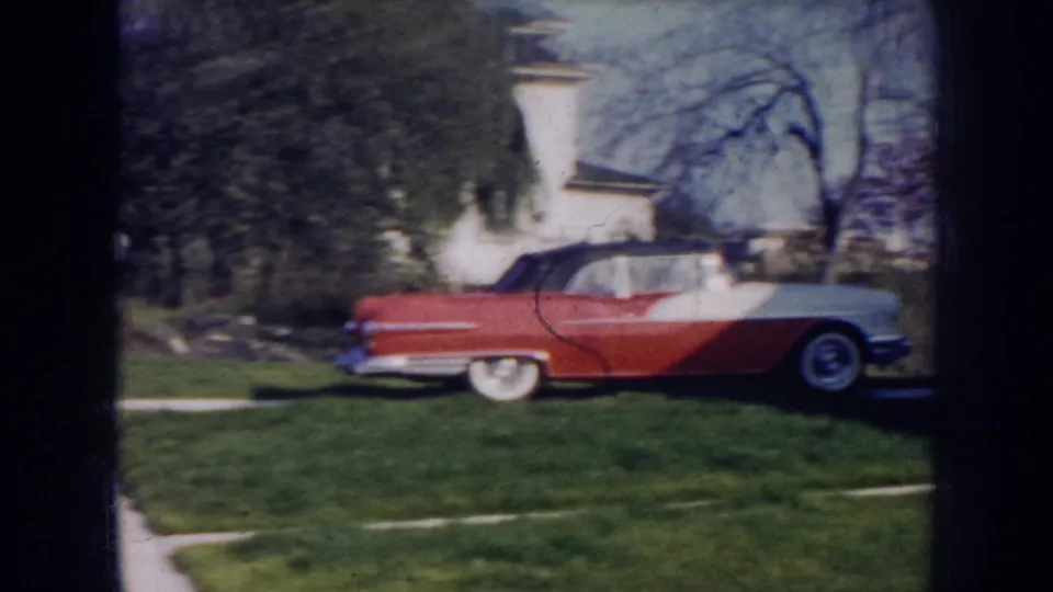 1956 Nebraska frame showing the side of a red-and-white convertible in a residential yard with bare trees behind it.