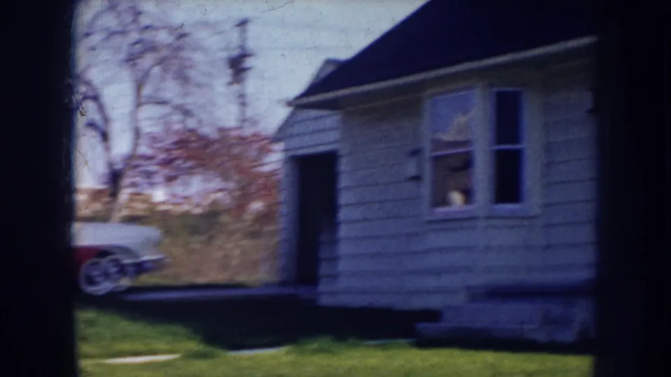 1956 Nebraska home-movie view of a light-sided house exterior with a partial car at the edge of the frame.