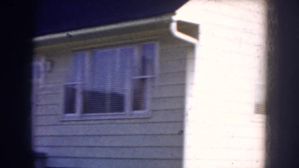 1956 Nebraska frame focused on a light-colored residential exterior with windows, siding, and bright sun on one wall.