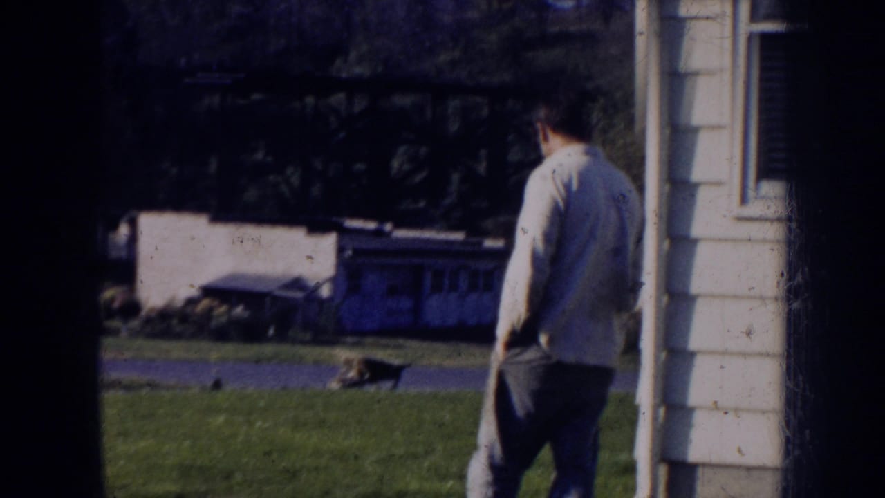 1956 Nebraska home-movie frame showing a man by a white-sided house, with lawn, street, and dark window framing visible.