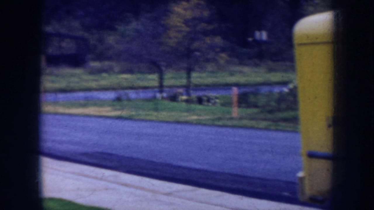 Nebraska residential street view from the 1956 8mm clip, with lawn, trees, pavement, and the edge of a yellow delivery truck.