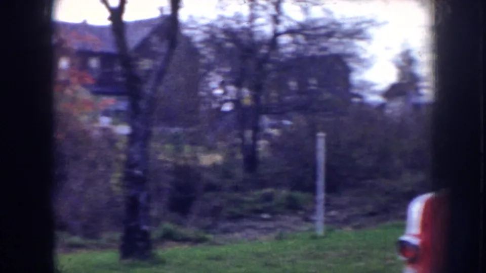 1956 Nebraska home-movie view of a quiet street with trees, lawns, houses in the distance, and a red vehicle edge.