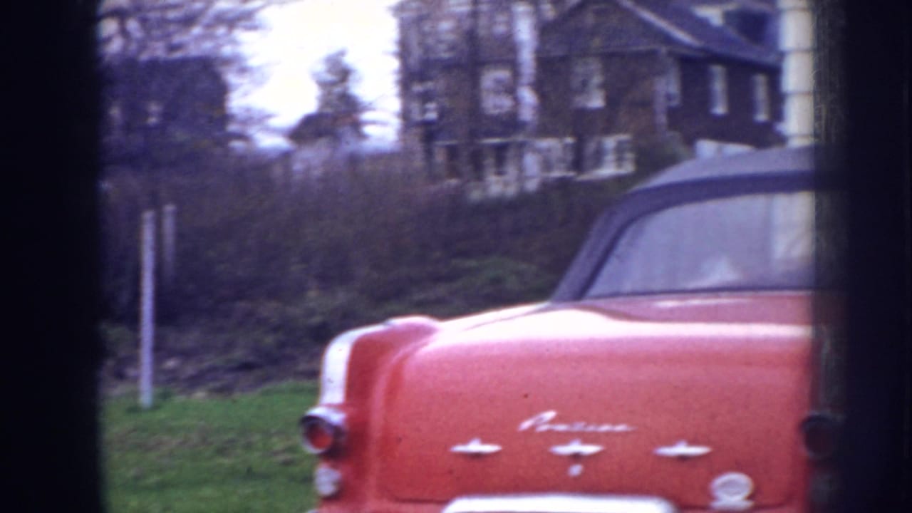 1956 Nebraska 8mm frame showing the rear of a red convertible near a suburban house and grass lawn.