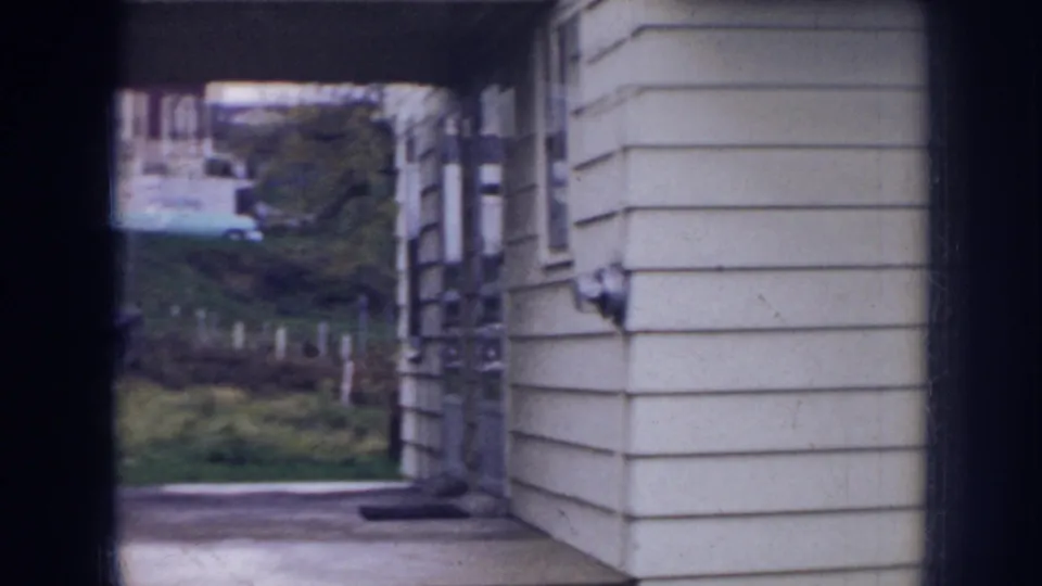 1956 Nebraska 8mm frame looking through a driveway entrance toward grass, shrubs, and neighboring homes.