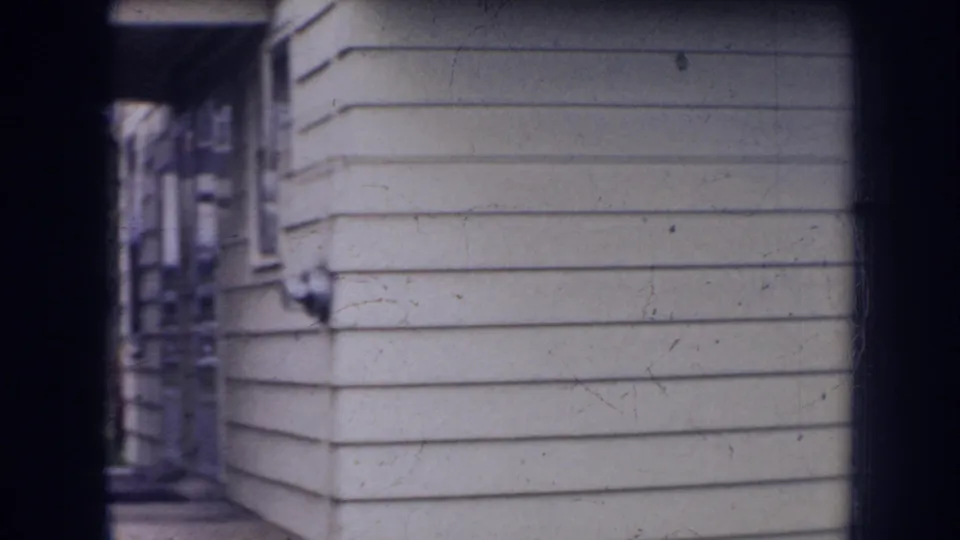 1956 Nebraska 8mm frame showing the side of a light-sided house with a narrow walkway and soft home-movie focus.