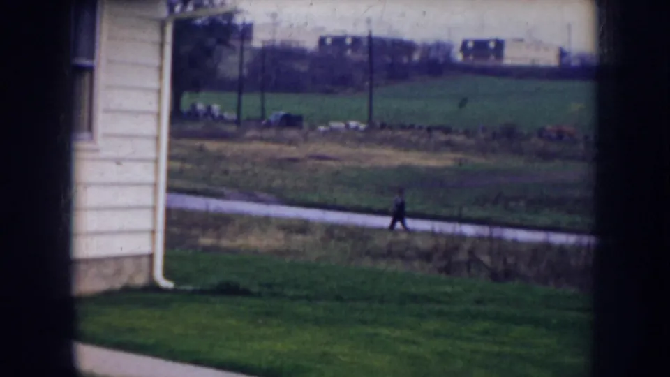 1956 Nebraska frame looking past a house corner toward a grassy area, path, fence, distant buildings, and one walking figure.