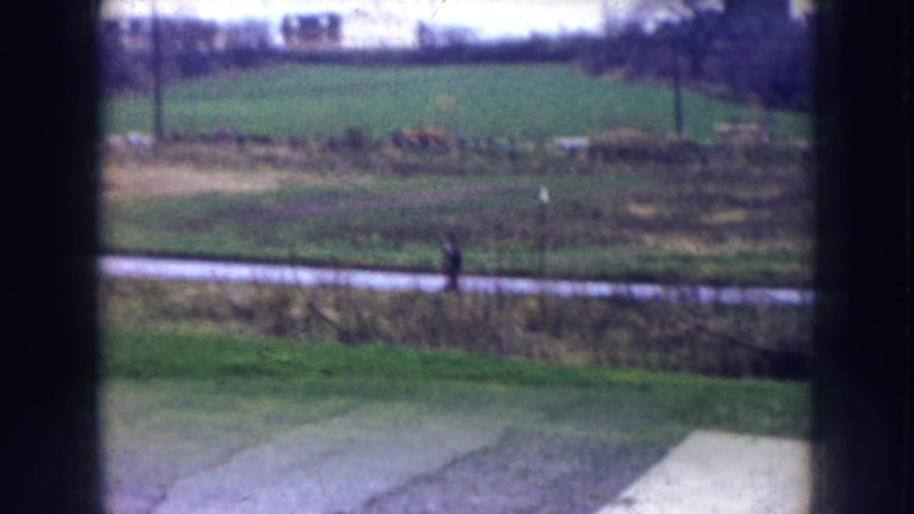 Distant pedestrian on a paved path with open fields seen through a window frame.