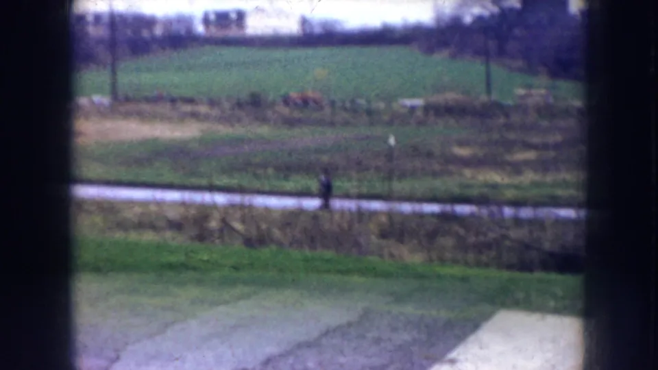 1956 Nebraska 8mm frame with a lone distant figure walking along a path beside open grassland, viewed through dark window framing.