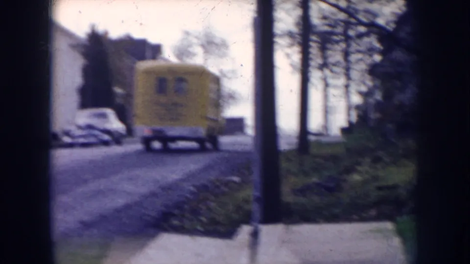 1956 Nebraska 8mm frame showing a yellow delivery-style truck on a residential road, with parked cars, houses, trees, and a utility pole nearby.