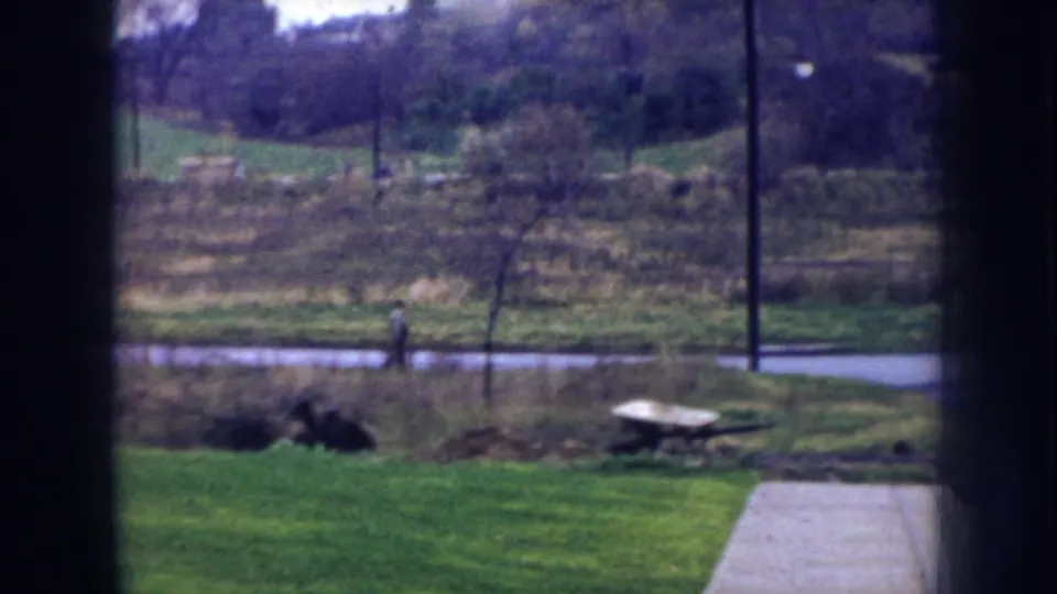 1956 Nebraska 8mm frame with a distant walker on a path, green lawn, open field, utility poles, and a wheelbarrow in the foreground.