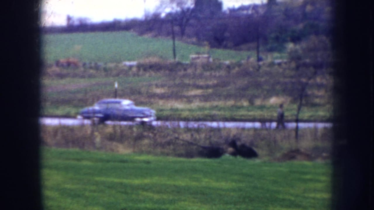 1956 Nebraska 8mm frame seen through a window, showing a blue car on a road beyond a green lawn and open fields.