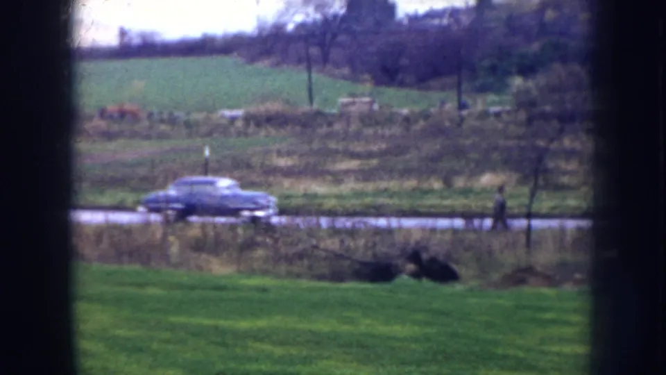 1956 Nebraska 8mm frame seen through a window, showing a blue car on a road beyond a green lawn and open fields.