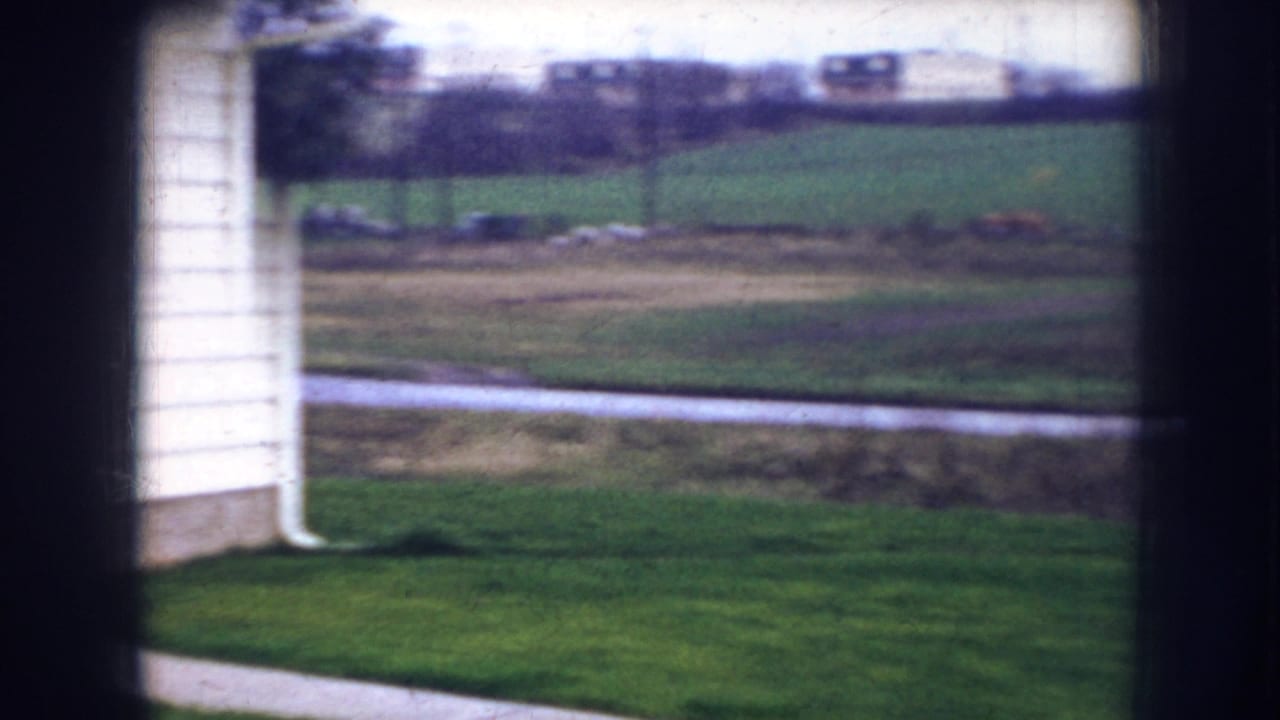 1956 Nebraska home-movie frame through a window, with pale siding, green lawn, road, ditch, and fields beyond.