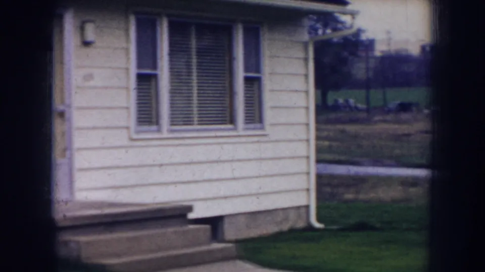1956 Nebraska frame showing a pale house exterior, small steps, window blinds, lawn, and distant field through a divided window.