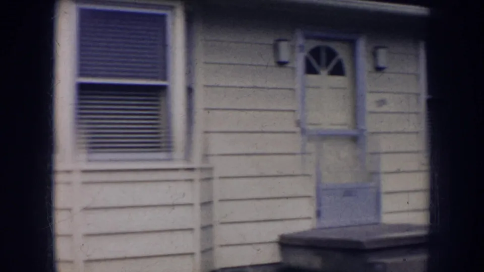 1956 Nebraska 8mm frame of a pale residential exterior with door, window blinds, small stoop, lawn, and road beyond.