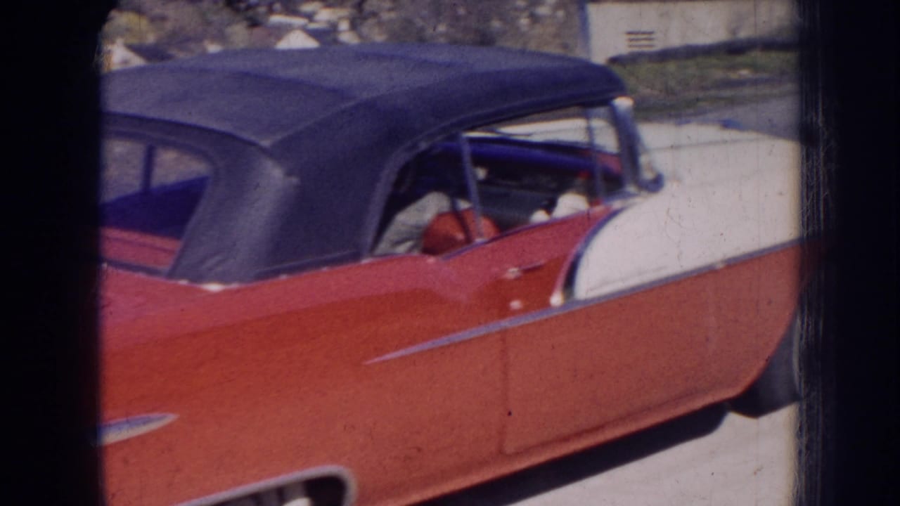 1956 Nebraska home-movie frame of a red-and-white convertible with a dark soft top, chrome trim, and indistinct people inside.