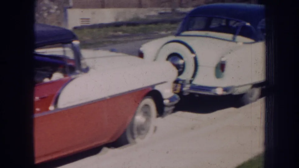 1956 Nebraska 8mm frame showing the rear of a white vintage car with dark roof and spare tire beside the red convertible.