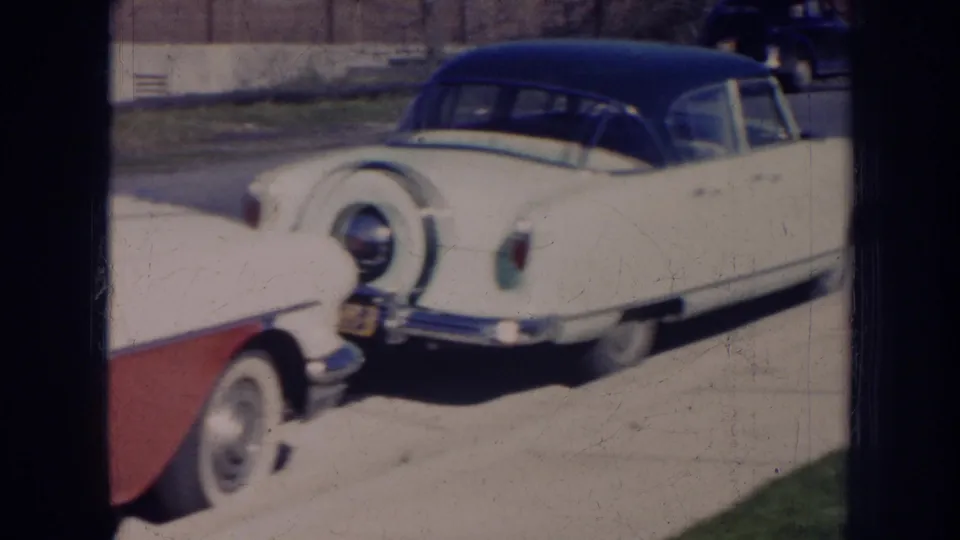 1956 Nebraska home-movie frame with a white mid-century car, dark roof, rear spare tire, and red convertible edge in view.