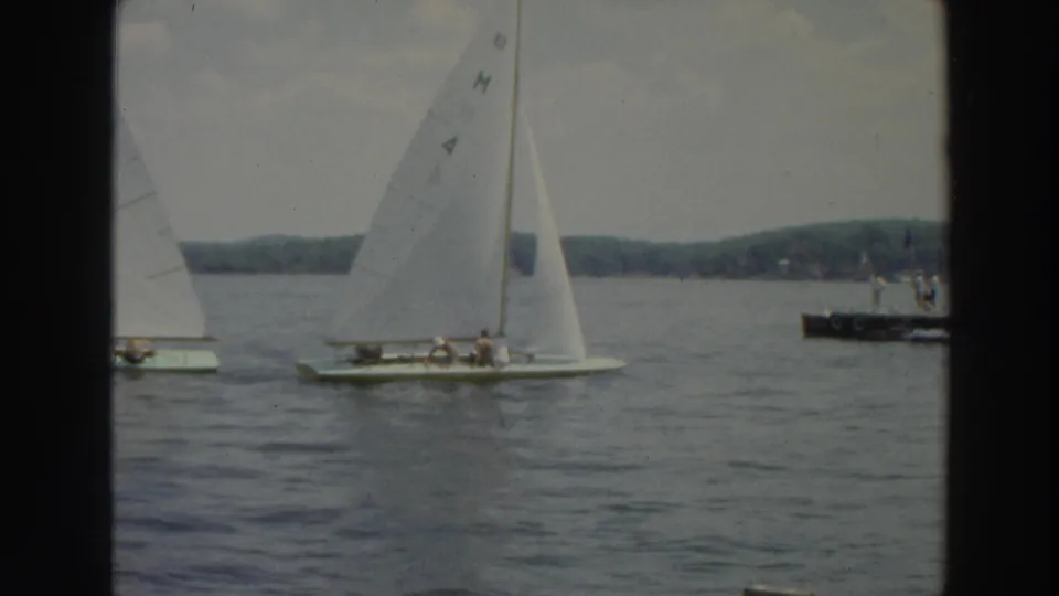 Wide 1966 Wisconsin lake view with white sailboats, small onboard figures, a dock, and wooded shoreline in the background.