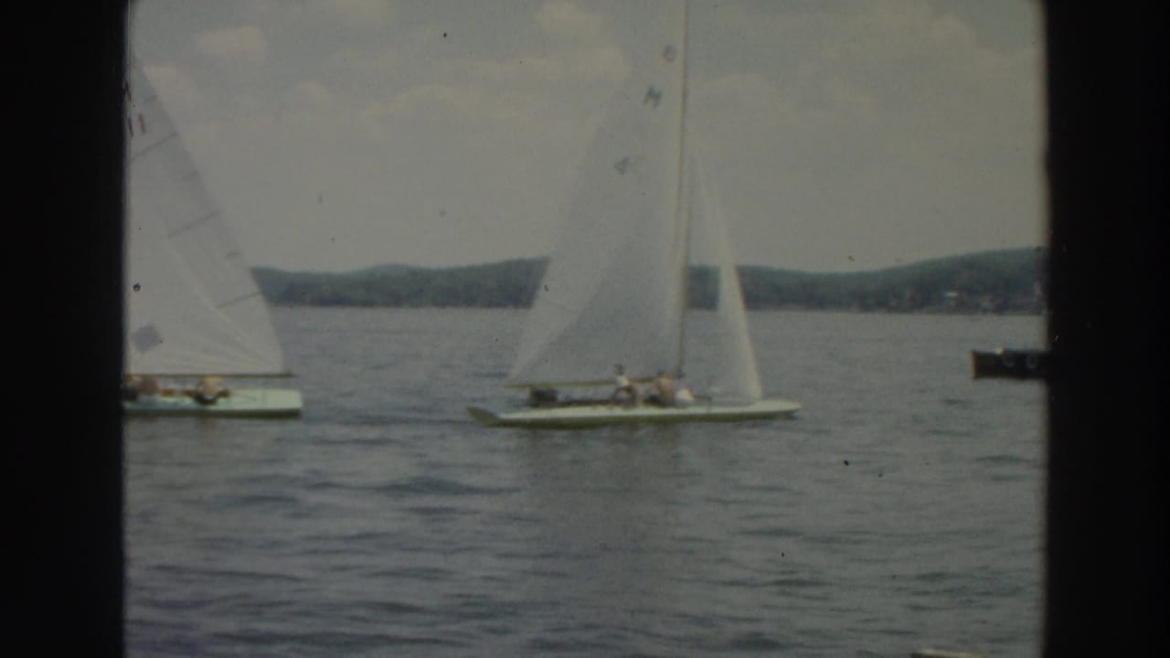 Two small sailboats with white sails move across a lake near a distant wooded shore.