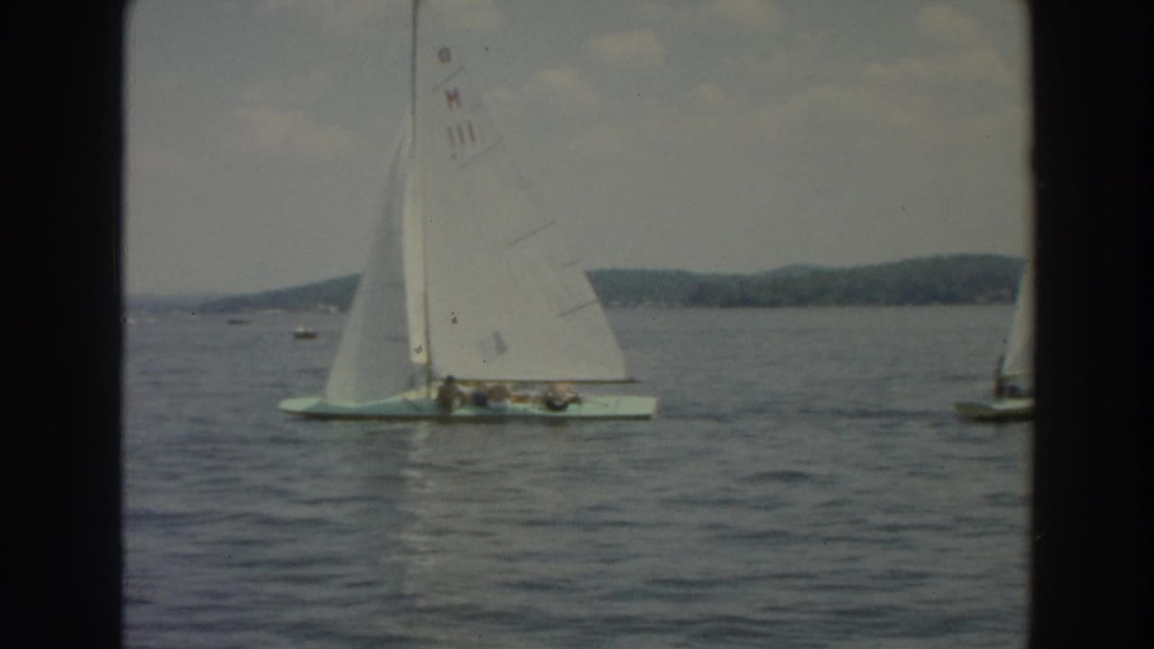 1966 Wisconsin 8mm frame of a white-sailed boat carrying several people on a lake, with distant shoreline behind it.