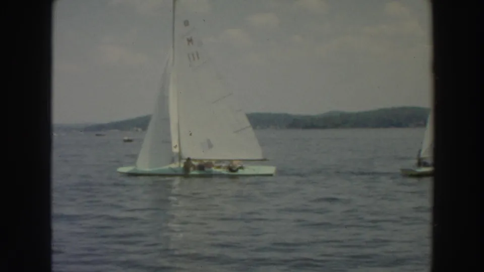 1966 Wisconsin 8mm frame of a white-sailed boat carrying several people on a lake, with distant shoreline behind it.