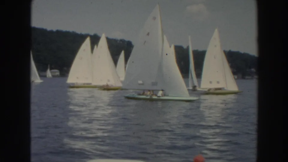 Small white-sailed boats pass in a loose line on a Wisconsin lake, with boaters faintly visible aboard.