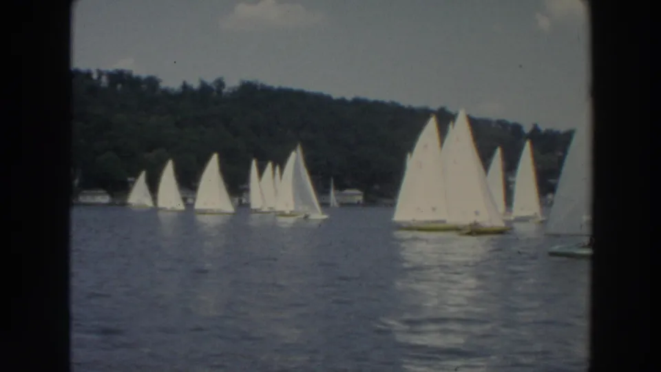 Wide 8mm view of small sailboats lined across a Wisconsin lake, with wooded shoreline and distant waterfront structures.