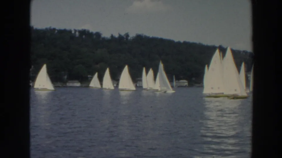 1966 Wisconsin 8mm frame showing multiple white sailboats spread across a calm lake below a dark tree-lined shore.