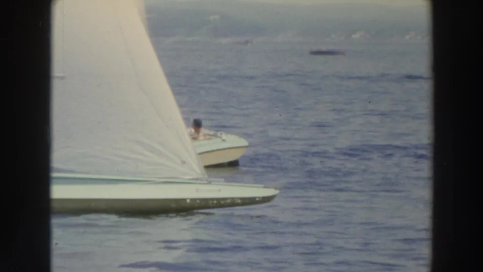 Close 1966 Wisconsin lake frame with a white-sailed boat crossing the foreground and a visible occupant in a nearby small boat.
