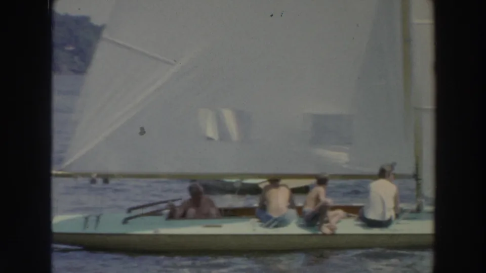 1966 Wisconsin home-movie frame with several people seated in a white sailboat under a large sail on lake water.