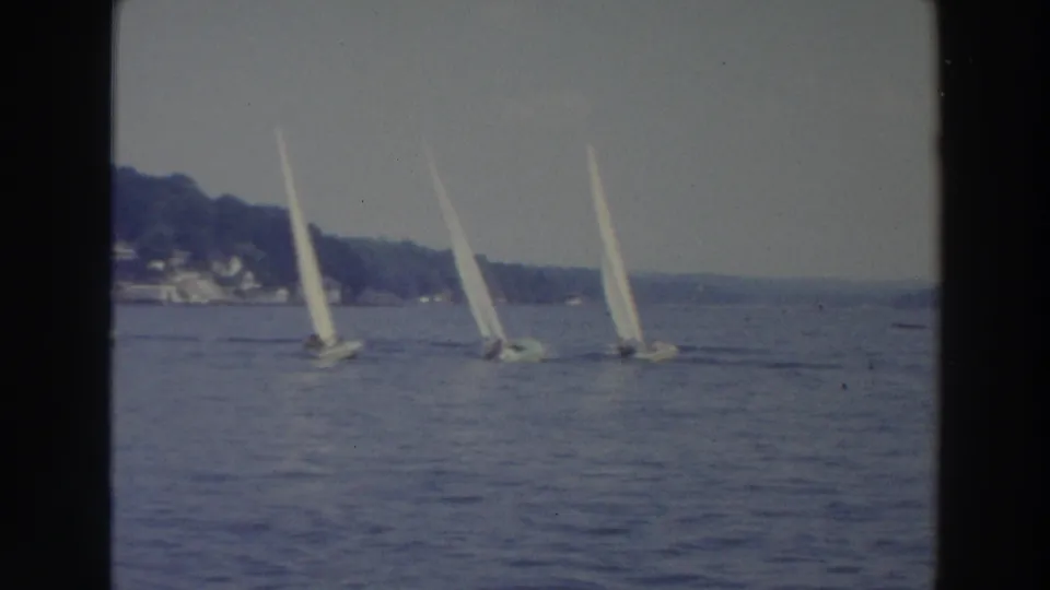 1966 Wisconsin home-movie frame with multiple white sailboats crossing a broad lake near a hazy, tree-lined shore.