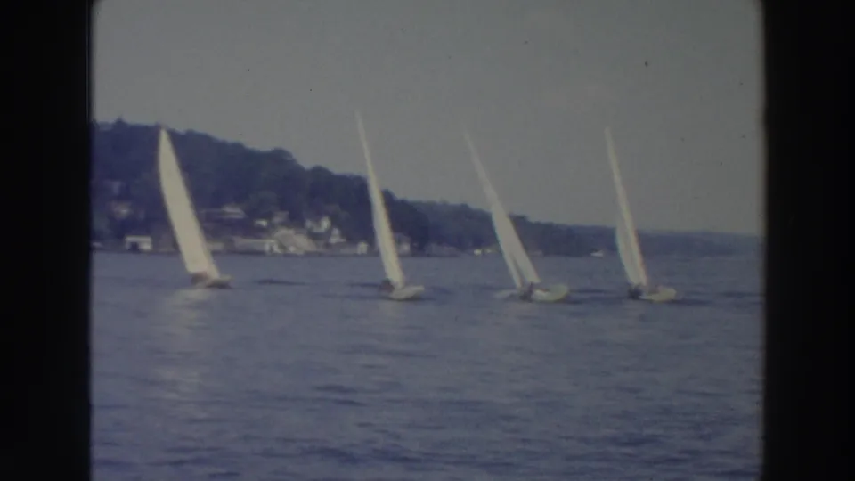 1966 Wisconsin 8mm frame of sailboats crossing a calm lake, with trees and distant shoreline structures in the background.