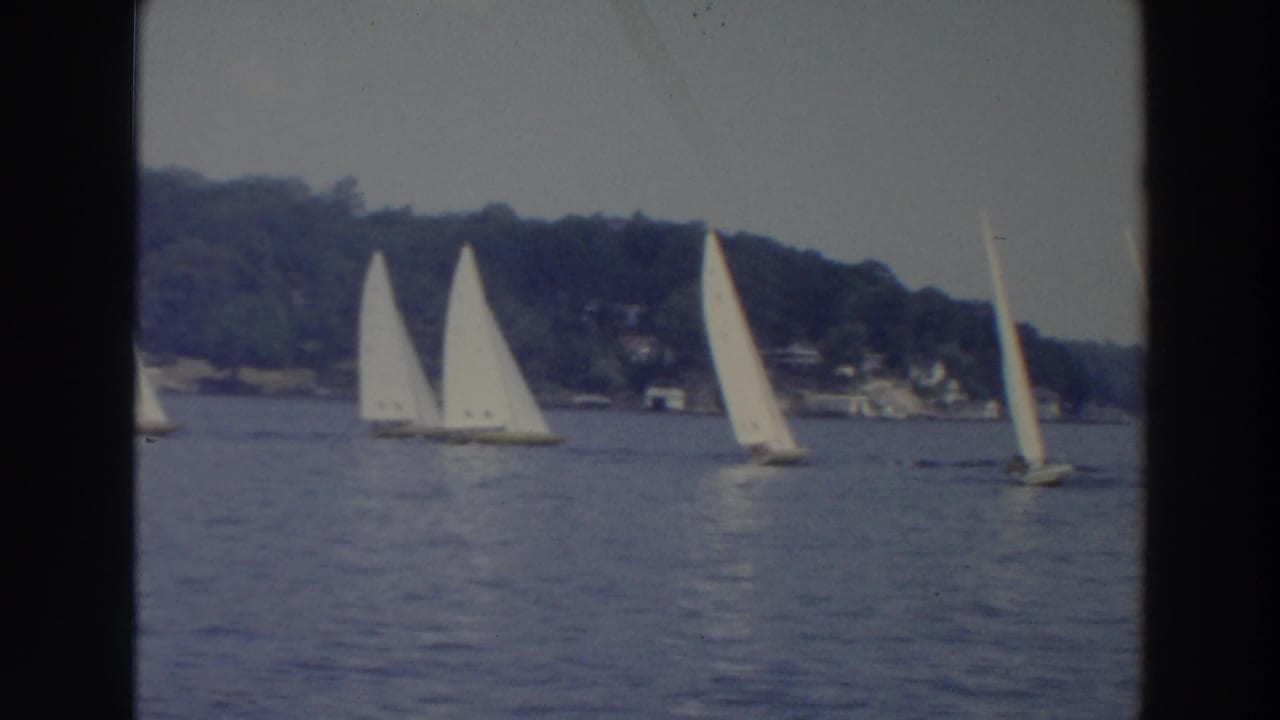 1966 Wisconsin 8mm view of multiple sailboats under white sails on a lake, with a wooded bank and small shore structures beyond.