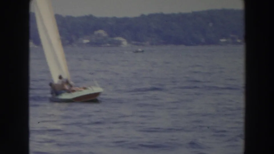 1966 Wisconsin lake footage frame with a small sailboat under a white sail crossing blue water near a tree-lined shoreline.