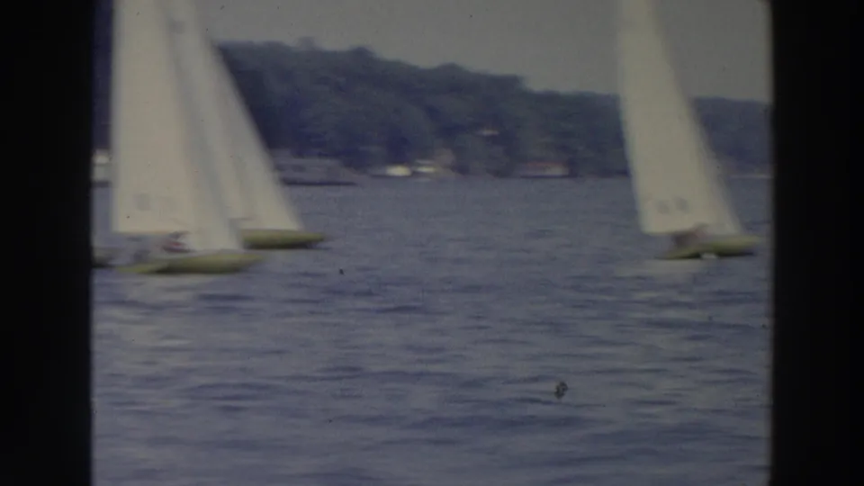 1966 Wisconsin 8mm frame showing two white-sailed boats crossing blue lake water below a wooded shoreline.