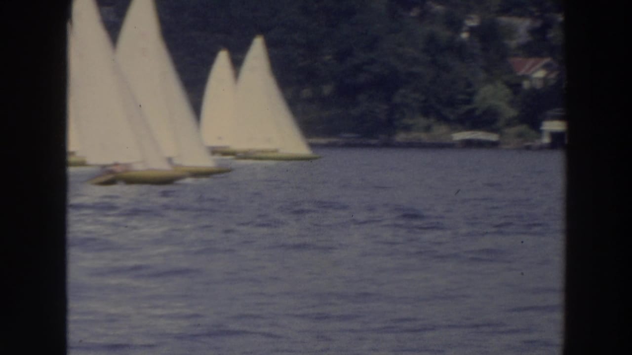 1966 Wisconsin 8mm frame showing small sailboats with white sails moving across rippled water near trees and distant houses.